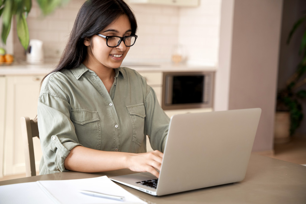 Smiling indian young adult woman wearing glasses typing on laptop computer working at home office sitting at table. Happy female professional freelancer student studying online using notebook pc.
